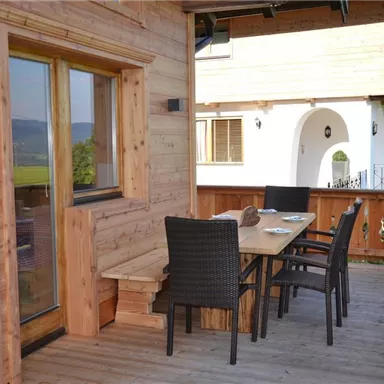 A cozy terrace with wooden paneling and a large table, surrounded by chairs. In the background, the house and the green landscape can be seen.