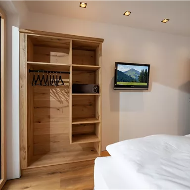 A modern bedroom with a wooden wardrobe and a small television on the wall. Large windows let in plenty of daylight and offer a view of the surroundings.