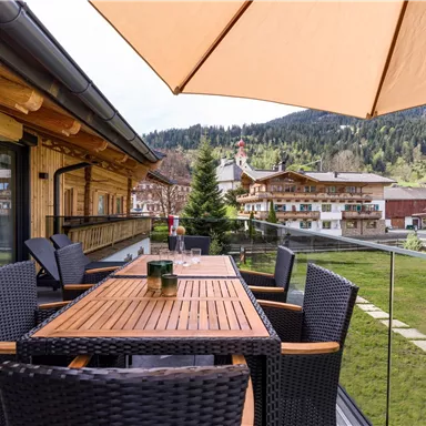 An elegant balcony with a wooden table and rattan chairs. In the background, picturesque mountains and wooden chalets can be seen.