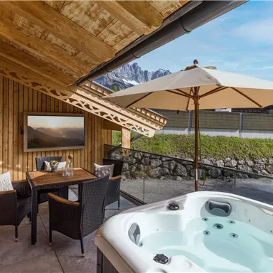A cozy outdoor area with a hot tub and a dining table under a sun umbrella. In the background, the mountains and a clear sky are visible.