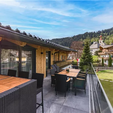 A modern terrace with wooden furniture and a view of green meadows and mountains. The surroundings are quiet and inviting.
