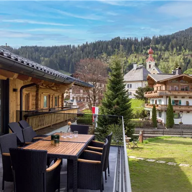 A cozy terrace with a wooden table and chairs, surrounded by a green meadow and houses in traditional architecture. In the background, trees and majestic mountains can be seen.