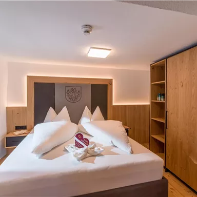 A modern hotel room with a large double bed and wooden details. The decor is bright and inviting, with a window and a wardrobe.
