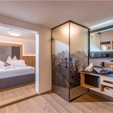 A modern room with a comfortable bed and elegant wooden elements. The bathroom is separated by a stylish glass wall.