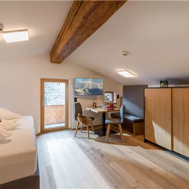 A cozy room with a large bed, a table, and chairs. It has windows with a view of the mountains and a warm wooden floor.