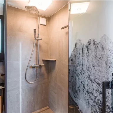 A modern shower with gray tiles and a glass partition. A stunning mountain motif can be seen on the wall.