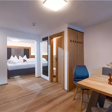 A modern hotel room with wooden flooring and elegant furniture. There is a dining area and a connection to another bedroom.