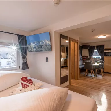 A cozy bedroom with two beds, a television, and large windows. The adjoining living area is modern and brightly furnished.