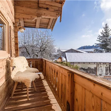 A cozy balcony with a wooden railing and two comfortable chairs. In the background, snow-covered trees and a clear sky can be seen.