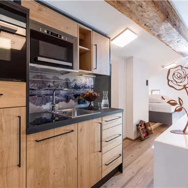 A modern kitchen with wooden cabinets and stainless steel appliances. In the background, a bedroom is visible with a decorative rose footstool.