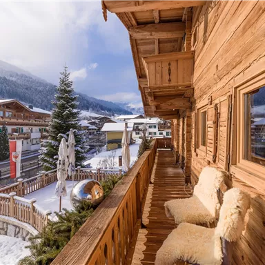 A cozy wooden porch with fur cushions and a view of a snowy landscape. In the background, traditional chalets and a Christmas tree can be seen.
