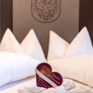 A cozy bed with white pillows and a heart-shaped welcome card in the center. The card reads "Welcome" and conveys an inviting atmosphere.