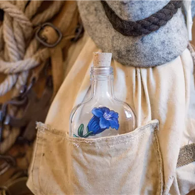 A bottle with a blue flower stands in a bag. In the background, ropes and other maritime objects can be seen.