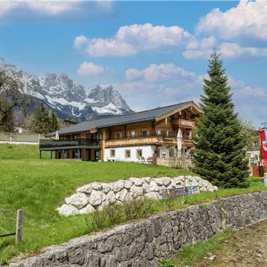 A cozy mountain cabin surrounded by green meadows and trees. In the background, impressive mountains and a clear sky can be seen.