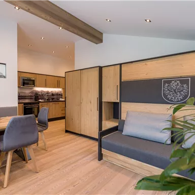 A modern living space with wooden details and a cozy sofa. The kitchen is open and well-equipped, with a large dining table nearby.
