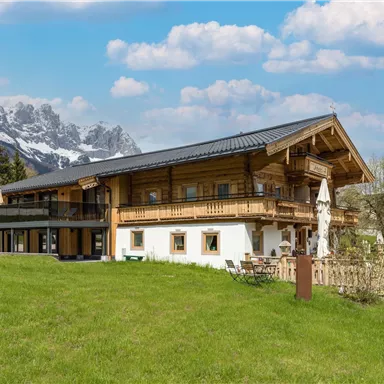 A beautiful wooden house with a modern design, surrounded by green meadows. In the background, majestic mountains and a blue sky can be seen.