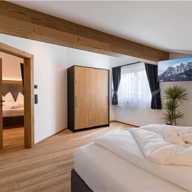 A modern bedroom with a wooden ceiling and large windows. It is bright and well equipped, with a television and a wardrobe.