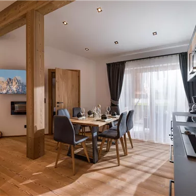 A modern living room with a wooden dining area and an open kitchen. Large windows provide plenty of daylight and a cozy atmosphere.