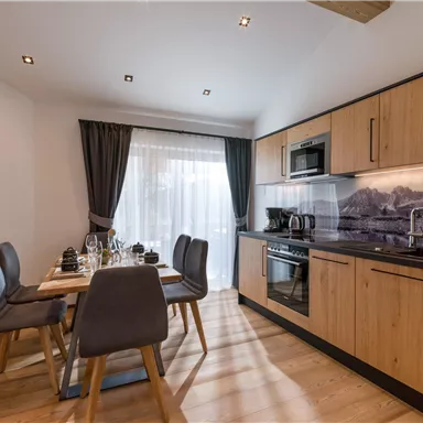A modern kitchen with wooden cabinets and a dining table. The windows are decorated with curtains, and the furnishings are well organized.