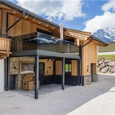 A modern chalet with wood and stone elements in a rural setting. In the background, mountains and a blue sky are visible.