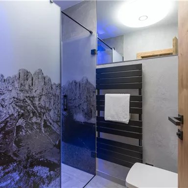 A modern bathroom with a glass shower and a stylish wall design. There is a towel holder and a bright ceiling light.