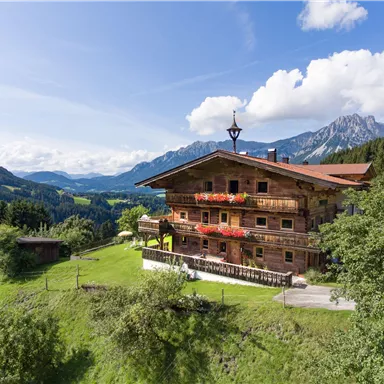Ein traditionelles Chalet in den Bergen mit schönen Blumenbalkonen. Die Umgebung ist landschaftlich reizvoll mit grünen Wiesen und Bergen im Hintergrund.