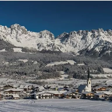 A picturesque winter landscape with snow-covered mountains and a small village. The scene features a clear blue sky with radiant sunshine.