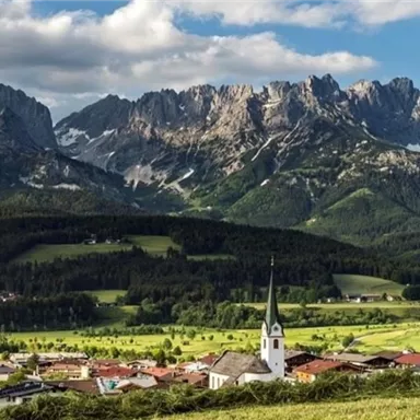 A picturesque landscape with high mountains in the background and a small village with a church in the foreground. The sky is partly cloudy and the meadows are green.