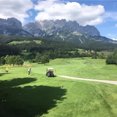 A picturesque golf course surrounded by mountains. The sky is blue and there is plenty of green grass.