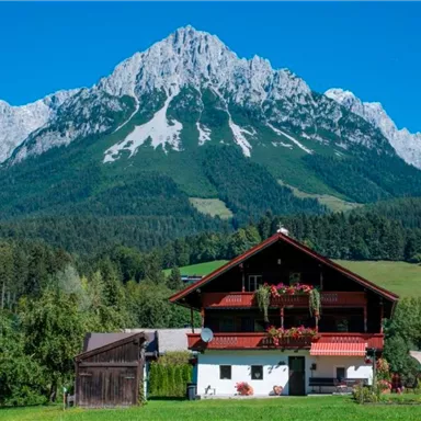 A picturesque alpine house in front of majestic mountains. The scene depicts a green landscape and a blue sky.
