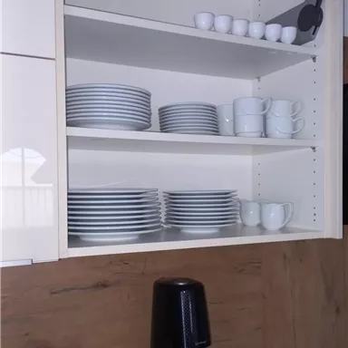 An organized kitchen cabinet with plates, cups, and small bowls. Below is a coffee machine and an information sheet.