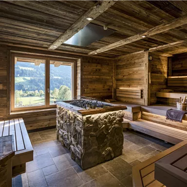 Ein moderner Wellnessbereich mit Holzverkleidung und großen Fenstern. Der Raum enthält eine Sauna mit einem steinernen Ofen und Holzliegen.