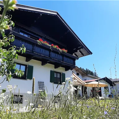 A traditional house with a balcony full of flowers and a large garden. The sun is shining in a clear blue sky.