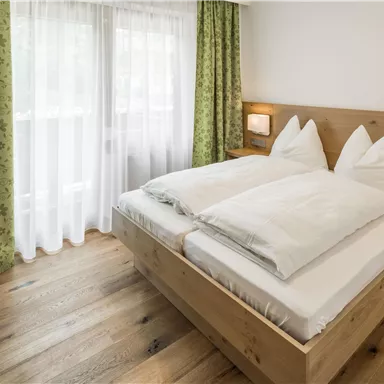 A bright bedroom with a large, comfortable bed and wooden floor. The windows are decorated with curtains made of green fabric.