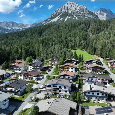 A picturesque mountain landscape with many wooden houses and a dense forest area. In the background, the majestic mountains of the Wilden Kaiser rise under a clear blue sky.
