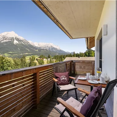 A beautiful balcony with a view of the mountains. Breakfast items are on the table and the sun is shining.
