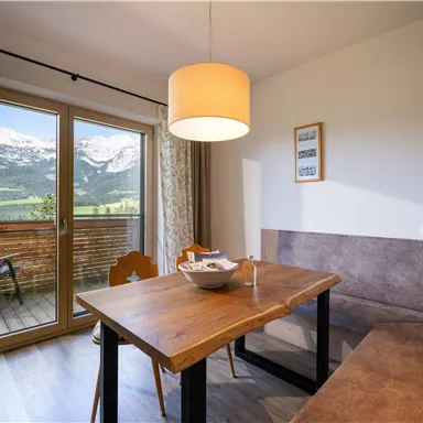 A cozy dining area with a wooden table and a bench. In the background are large windows that offer a beautiful view of the mountains.