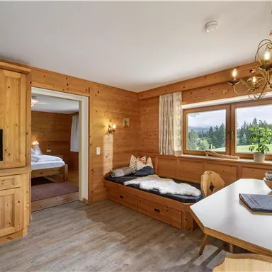 A cozy living area made of wood featuring a table and a sofa. Large windows provide a view of the green landscape outside.