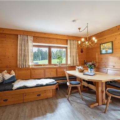 A cozy room with wooden panelled walls, a dining table, and a seating area. Large windows offer a view of the nature outside.