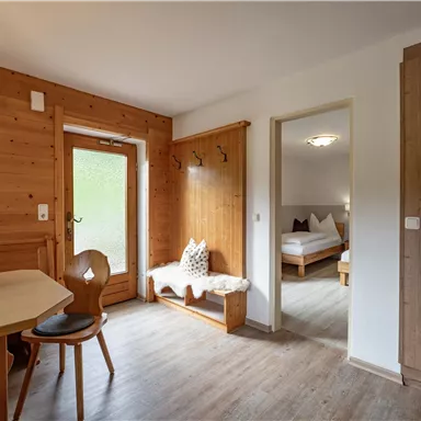 A cozy room with wooden paneling, a table, and chairs. In the background, an entrance and a bedroom can be seen.