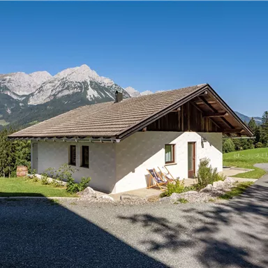 A comfortable house in the mountains, surrounded by green meadows. In the background, you can see majestic mountains under a clear sky.