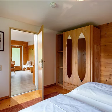 A cozy bedroom with a double bed and a wooden wardrobe. In the background, another room with natural light can be seen.