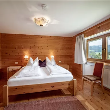 A cozy bedroom with wood paneling. There is a double bed, a chair, and a window with a view of the landscape.