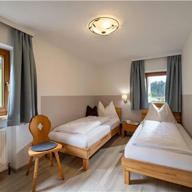 A simple room with two single beds, a chair, and a window overlooking the outdoors. The walls are painted light and the windows have curtains.