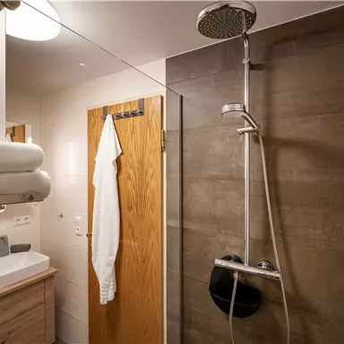 A modern bathroom with a rain shower and a wooden door. The sink is designed simply and elegantly, next to a towel and a bathrobe.
