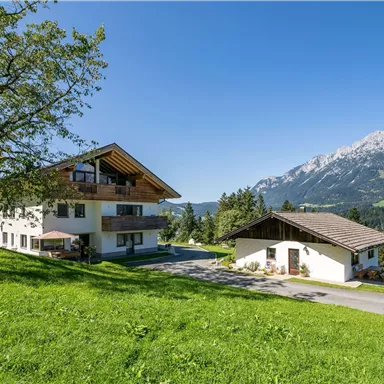 A picturesque landscape with a large house and a smaller building. In the background, majestic mountains and a clear blue sky can be seen.