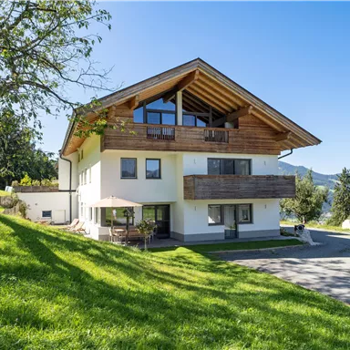 A modern house with a beautiful wooden roof and large windows. It is situated in a green setting with a view of the mountains.