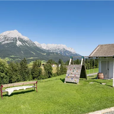 An idyllic landscape with a meadow, a climbing frame, and a seating area. In the background, majestic mountains rise under a clear blue sky.