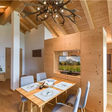 A bright and modern dining room with wooden decor and a large table. In the background, there is a television displaying a mountain panorama.
