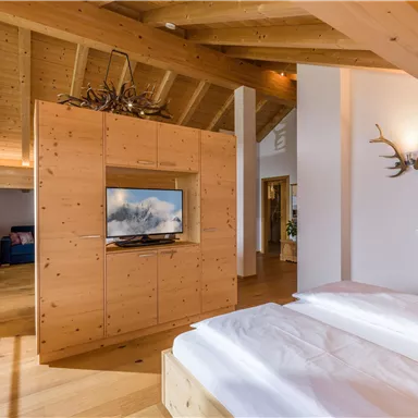A modern bedroom with wooden ceilings and a cozy wooden wardrobe. A television is built into the furniture, and there are large windows that let in plenty of light.